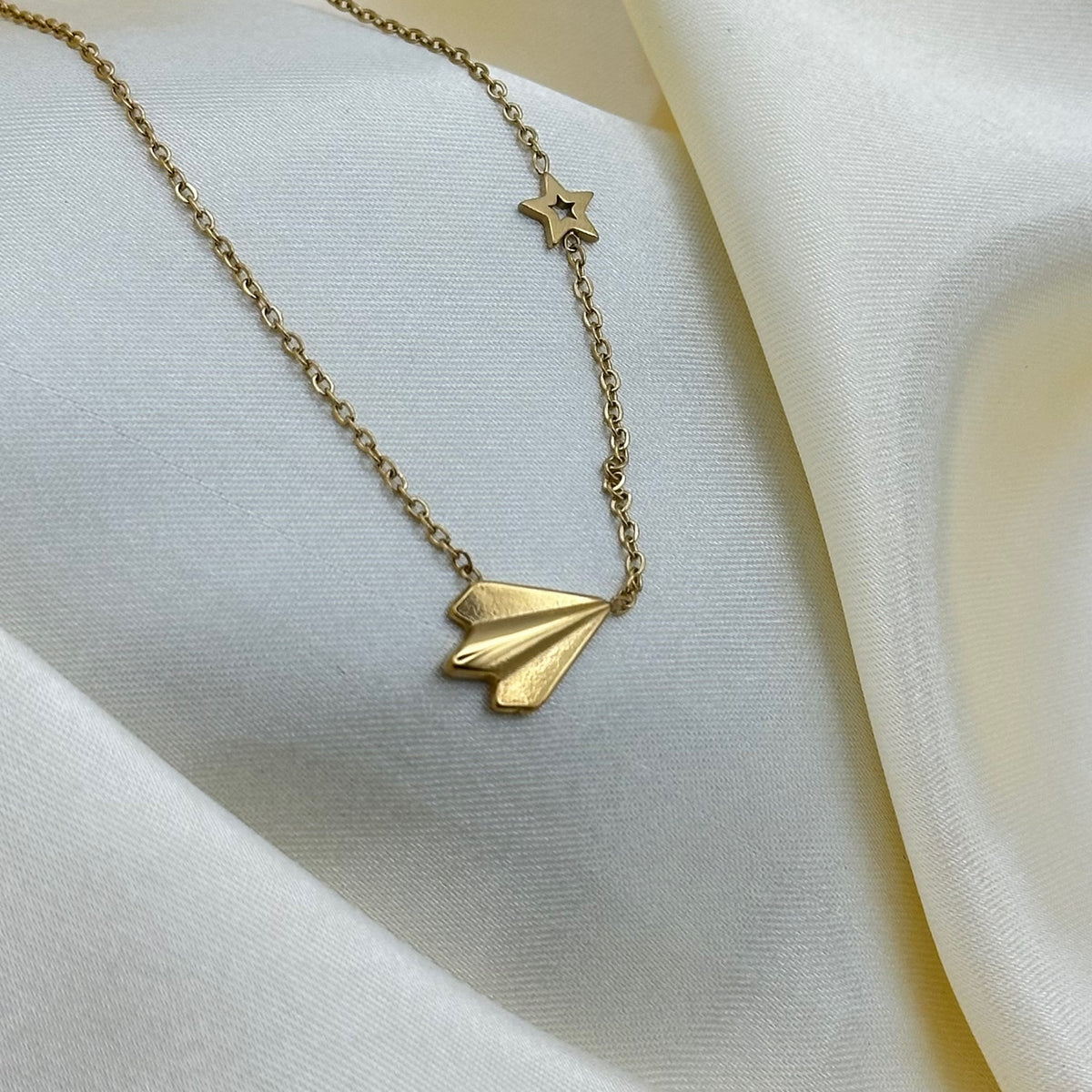 A gold origami paper plane pendant and star charm on a fine chain, resting on draped cream satin fabric.