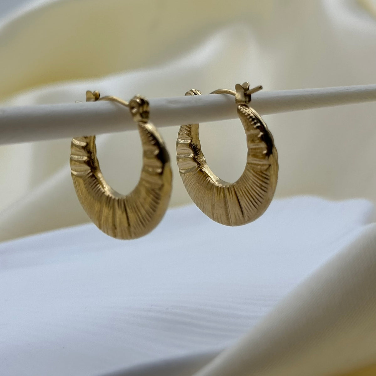 A pair of textured gold hoop earrings hanging from a white display rod against a soft cream-colored fabric background.