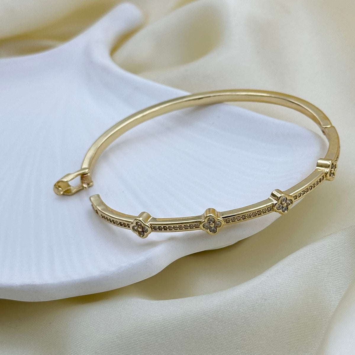 A gold-plated bangle bracelet with small clear crystal accents, displayed on a soft white textured surface with draped cream fabric in the background.