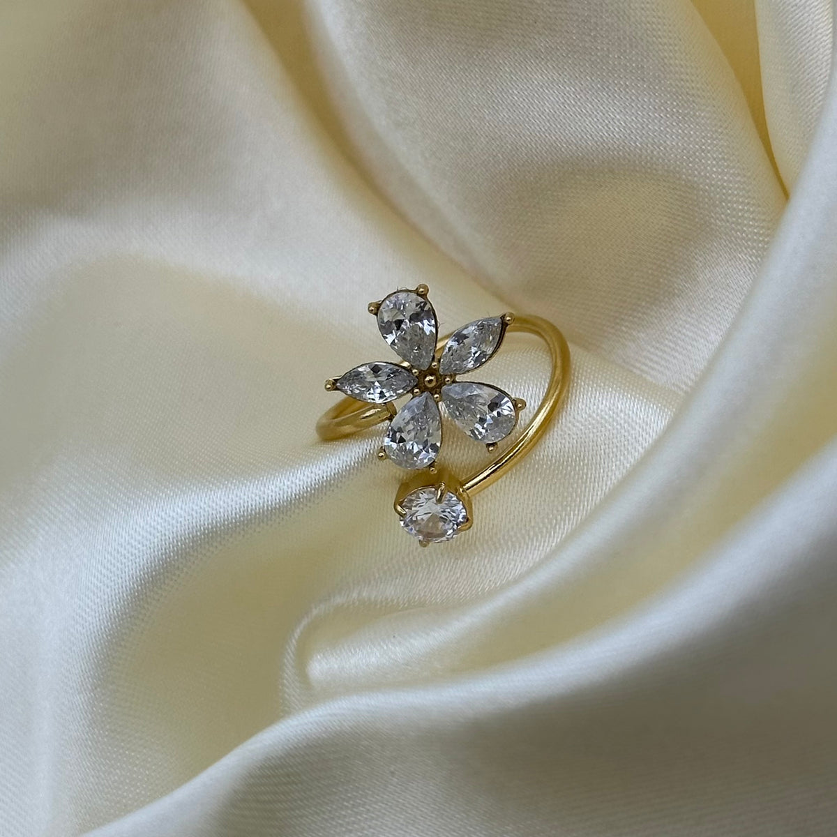 A gold flower-shaped ring with clear marquise and heart gemstones, displayed on draped cream-colored satin fabric.