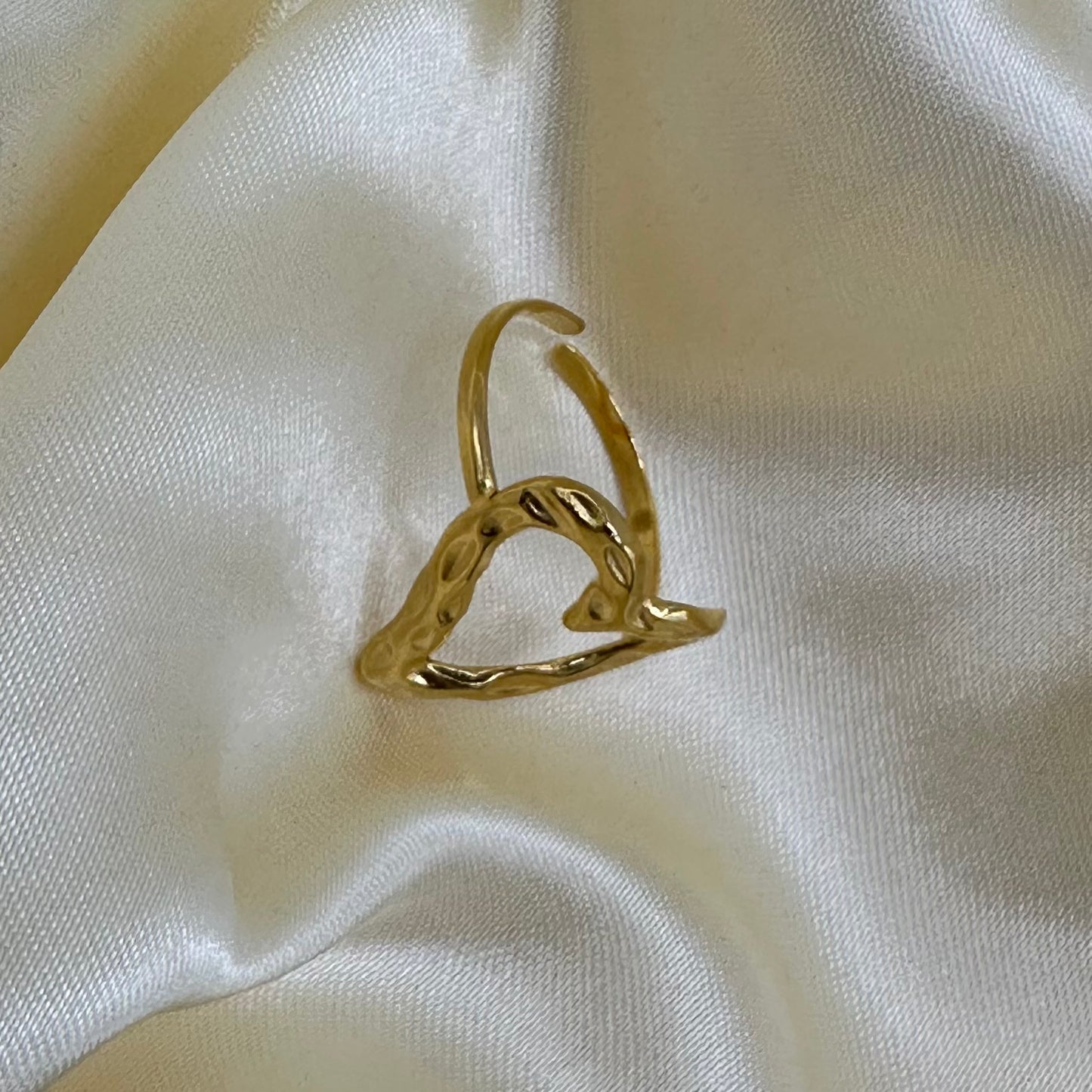 A hammered gold heart-shaped adjustable ring standing upright on draped white satin fabric.