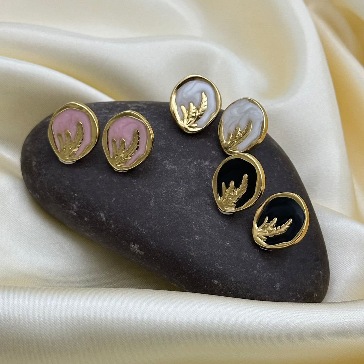 Three pairs of gold oval stud earrings with a raised botanical branch motif on pink, white and black enamel, arranged on a dark stone over cream satin fabric.