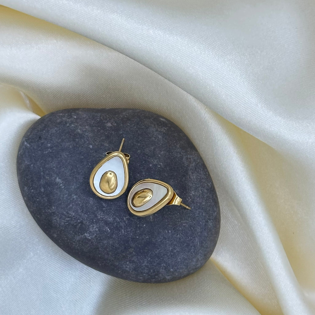 A pair of gold avocado-shaped stud earrings with white enamel and a polished gold pit centre, displayed on a dark stone over cream satin fabric.
