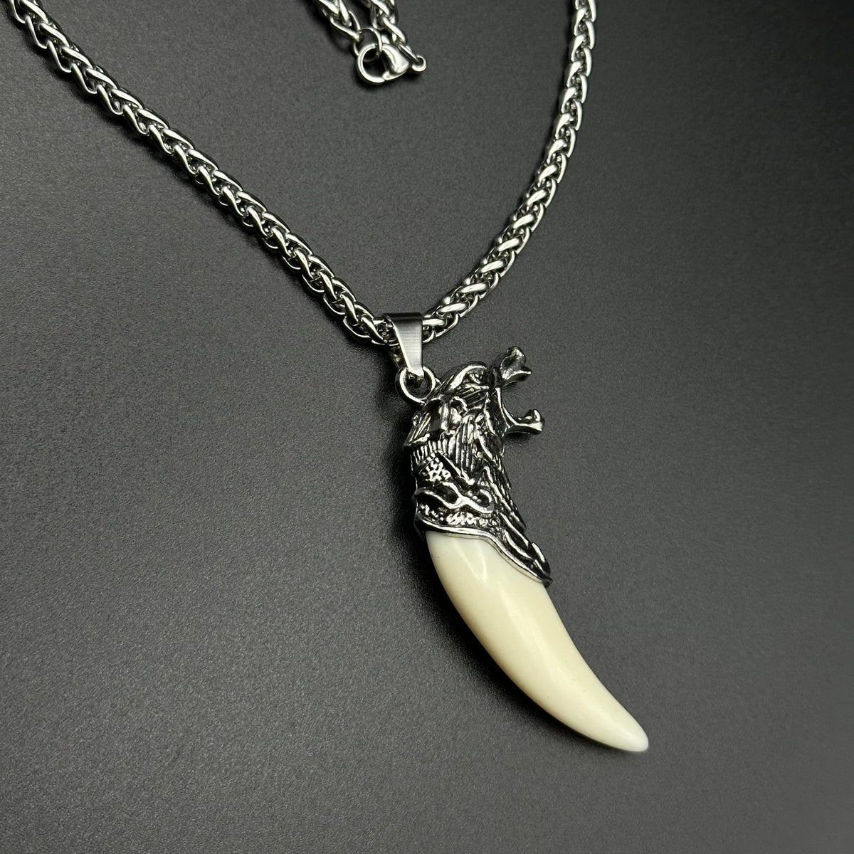 A silver wolf-head pendant with a curved cream fang on a braided silver chain, photographed against a dark grey background.