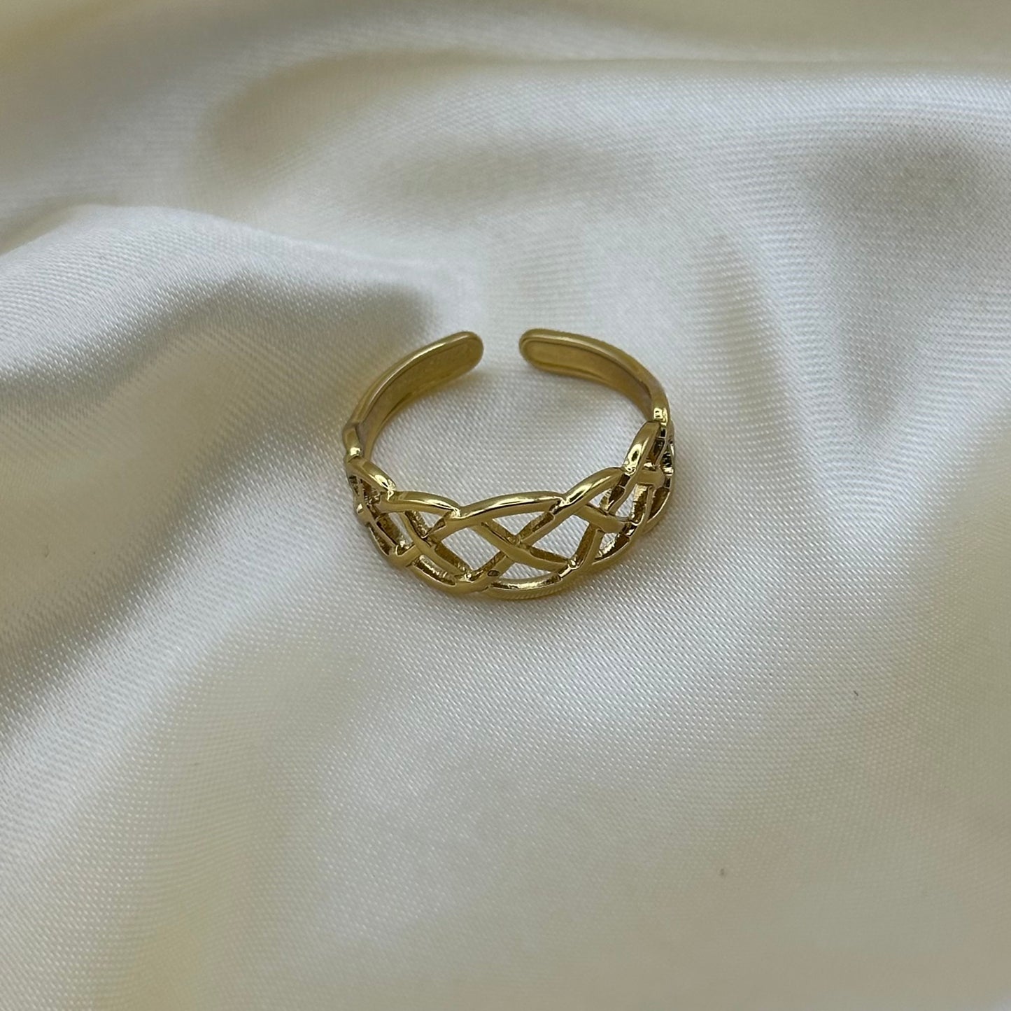 A gold-toned open cuff ring with an intricate woven or braided link pattern, resting on a draped cream-colored fabric background.