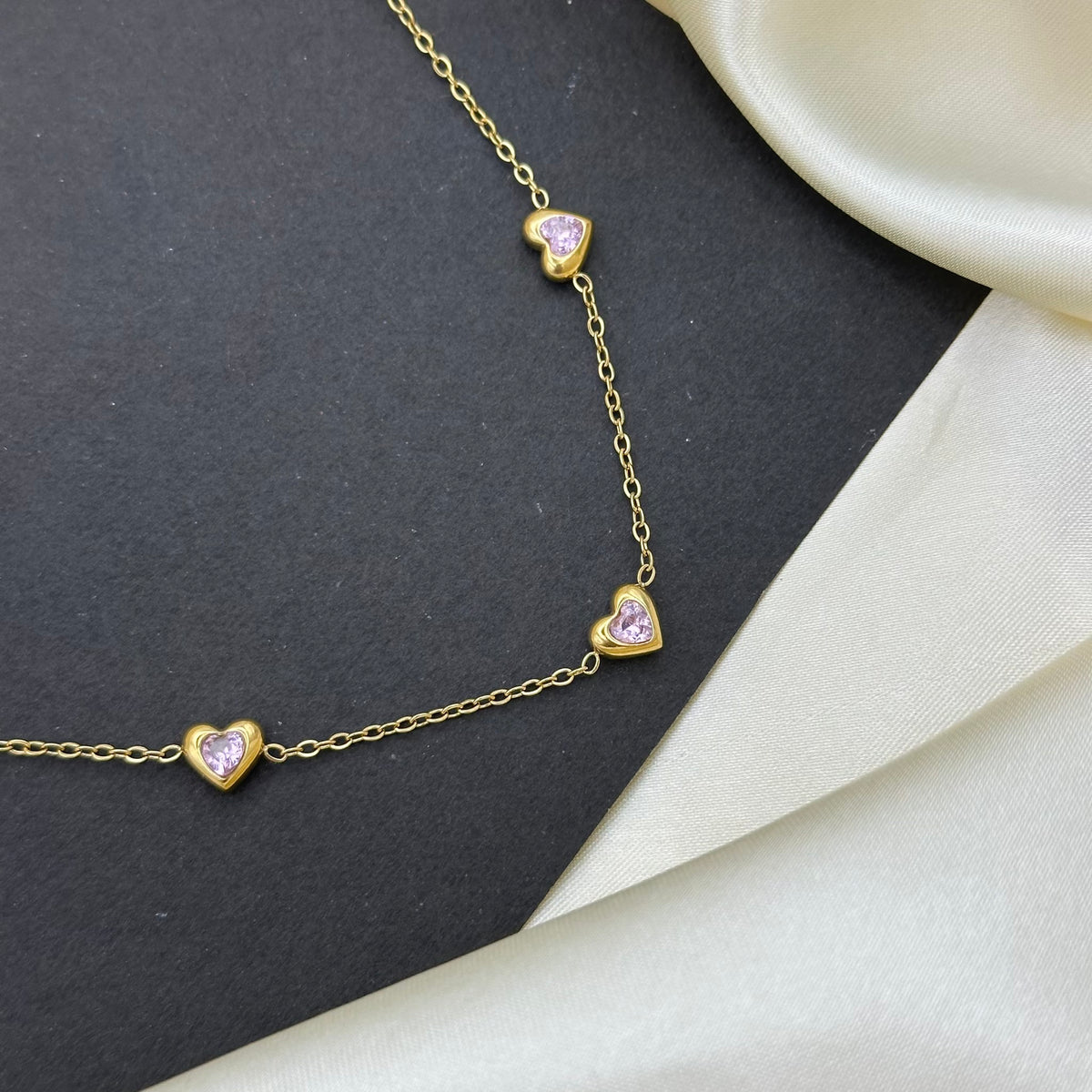 A delicate gold chain necklace featuring small heart-shaped charms with light pink inlays, styled on a dark gray textured surface with cream-colored satin fabric.