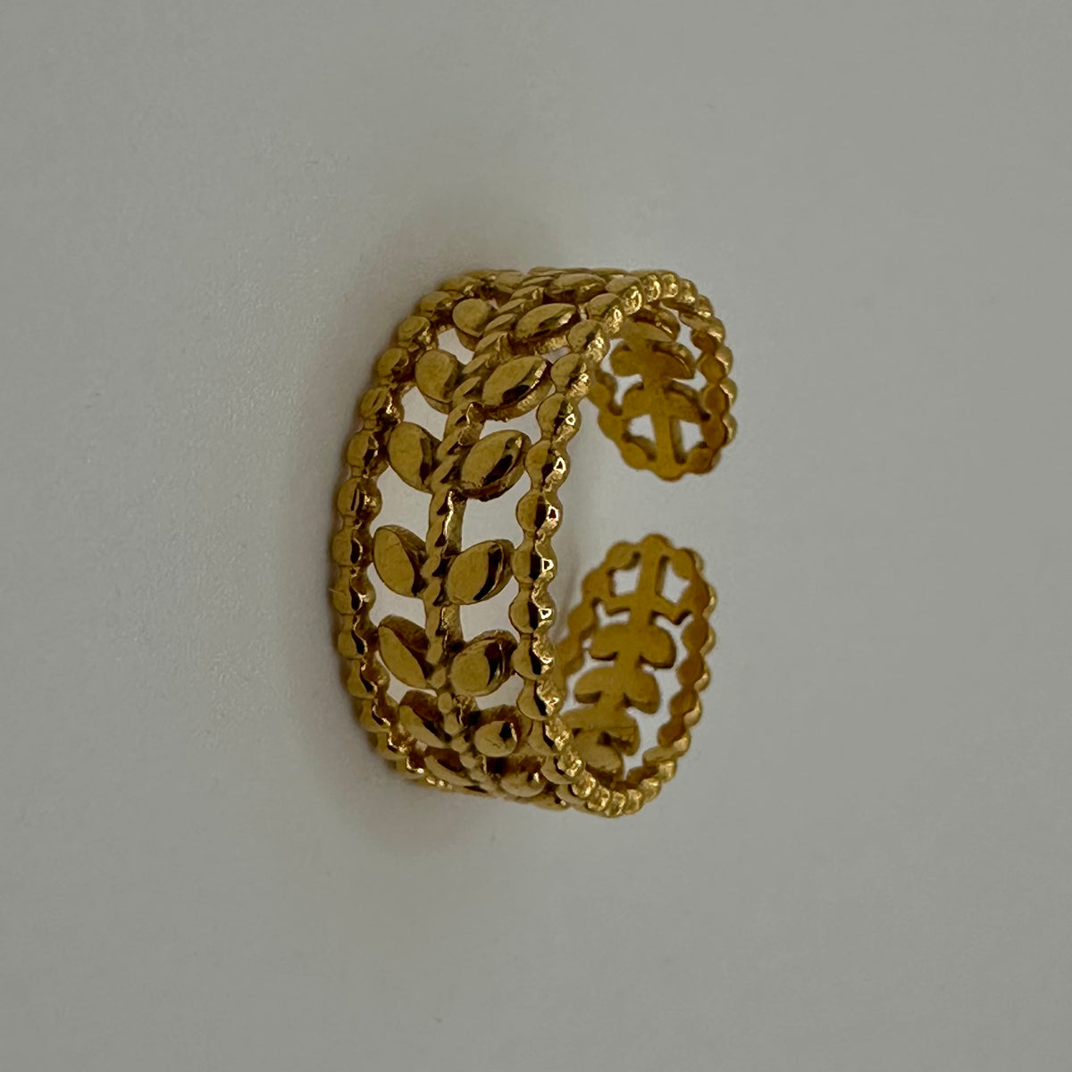 A gold-colored open cuff ring with a detailed filigree leaf pattern, displayed on a neutral light gray background.