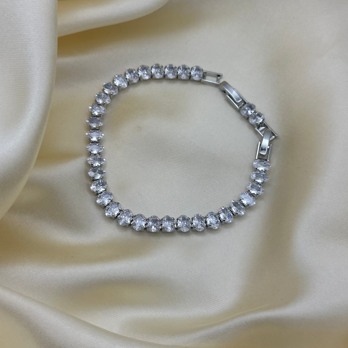 A silver tennis bracelet with clear oval-cut stones, displayed on a draped cream satin fabric background.