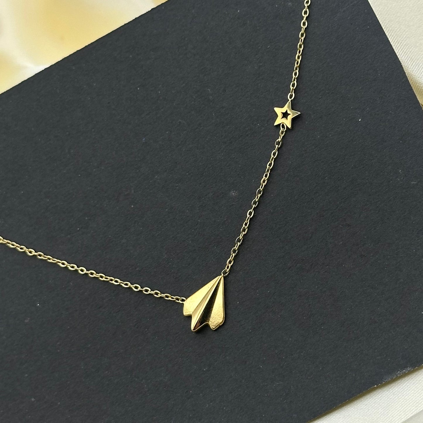 A gold paper plane pendant with a small star charm on a fine gold chain, styled on a dark grey card.