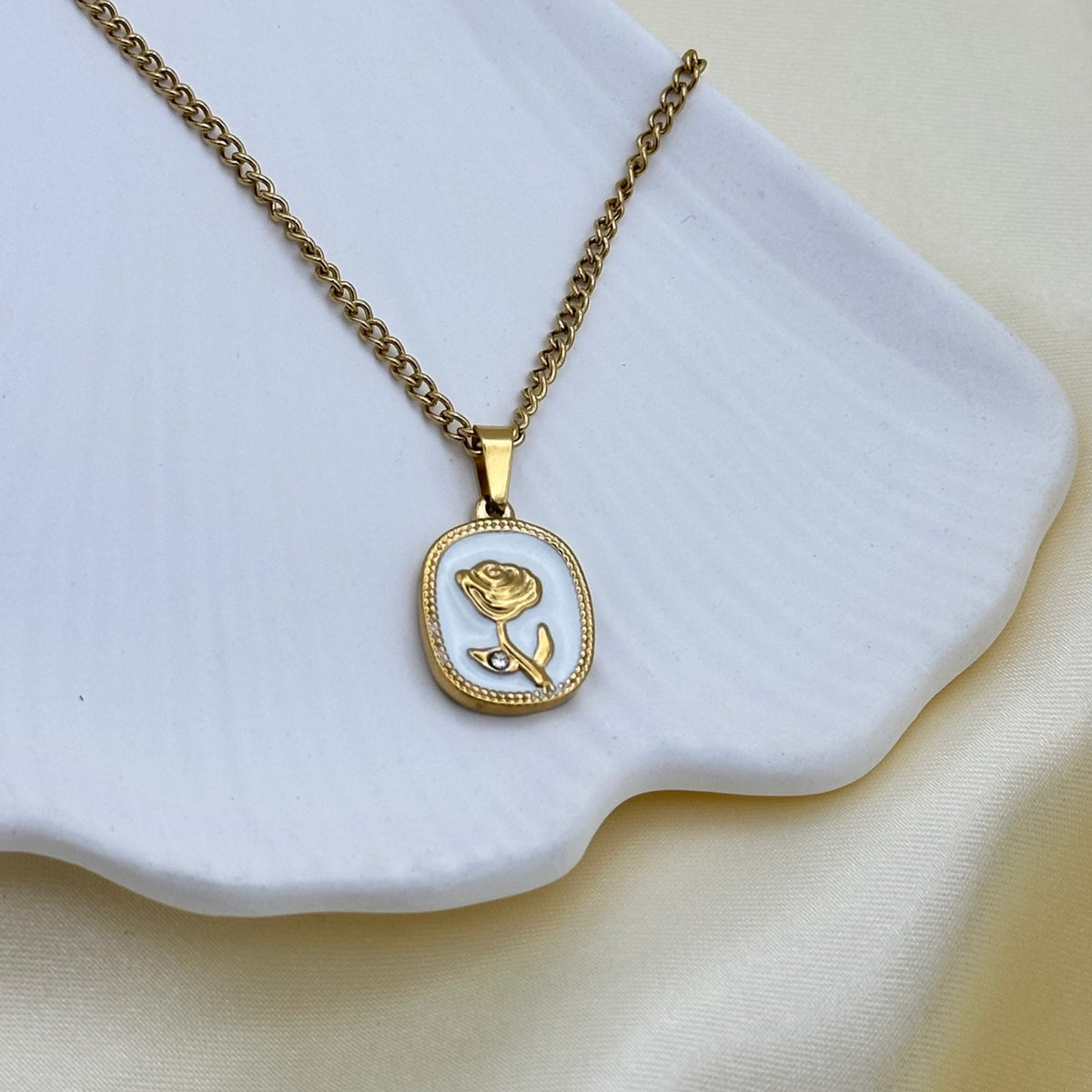 A gold cushion-shaped pendant with a white enamel face and a raised gold rose set with a tiny crystal, on a curb chain over a white ceramic dish.