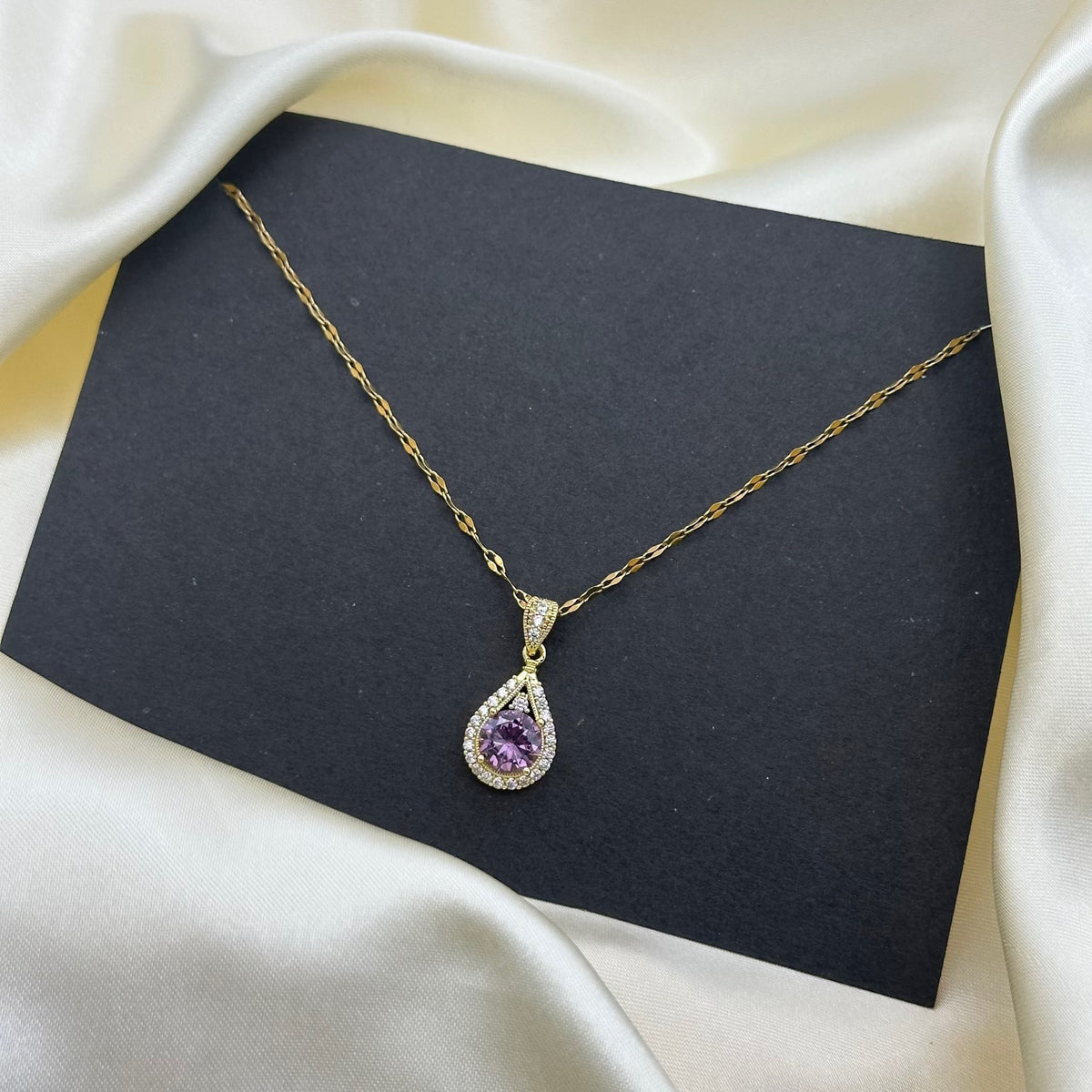 A purple teardrop crystal pendant with a clear stone halo on a fine gold chain, styled on a dark grey card.