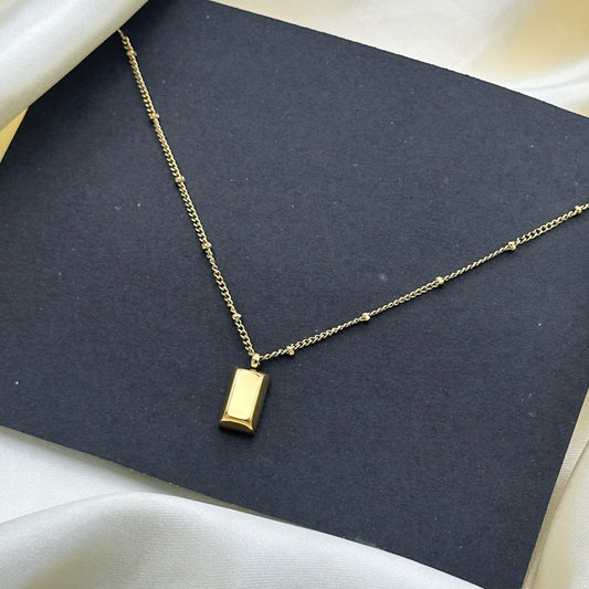 A polished gold rectangular bar pendant on a delicate station chain, styled on a dark grey card over cream satin.