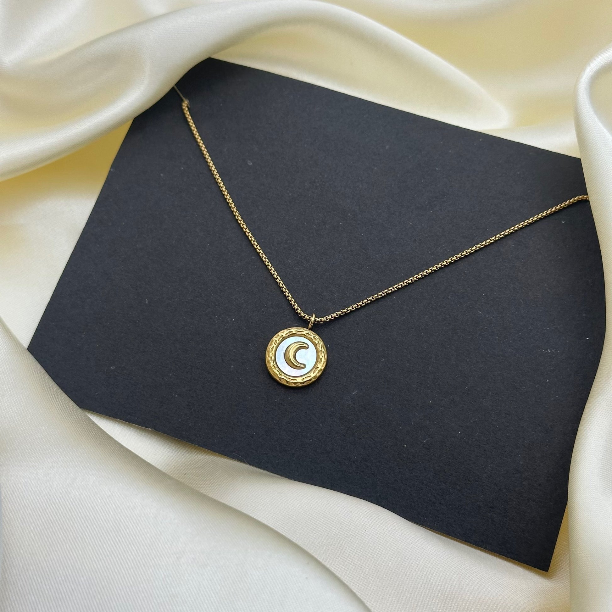 A small gold pendant with a white enamel face and a raised gold crescent moon on a fine chain, styled on a dark grey card.