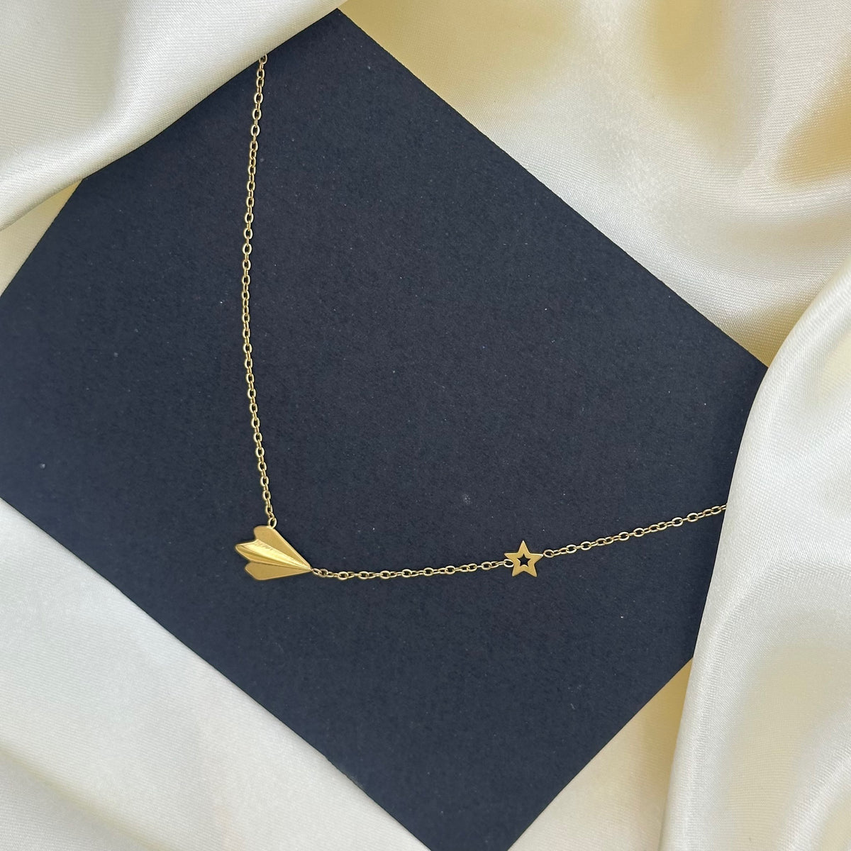 A delicate gold necklace with an origami paper plane pendant and star accent, displayed on a dark grey card over cream satin.