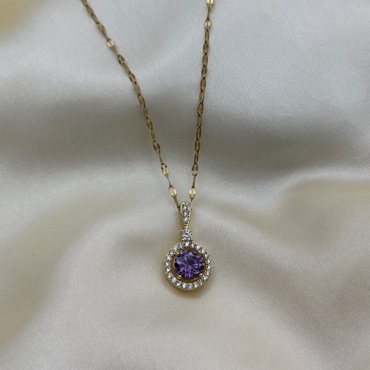A round purple crystal halo pendant on a textured gold chain, shown against soft cream satin.