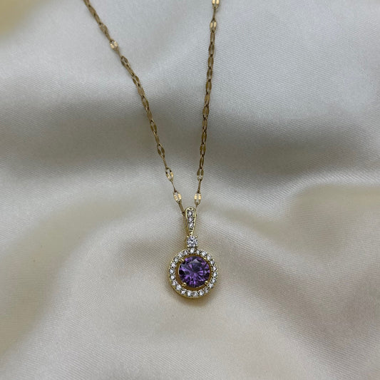 A round purple crystal halo pendant on a textured gold chain, shown against soft cream satin.