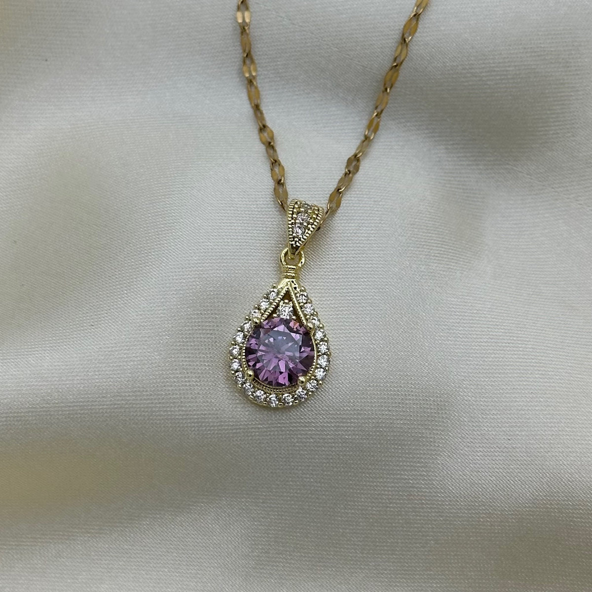 A gold teardrop pendant set with a purple crystal surrounded by a halo of clear pave stones, on a textured gold chain over cream satin.