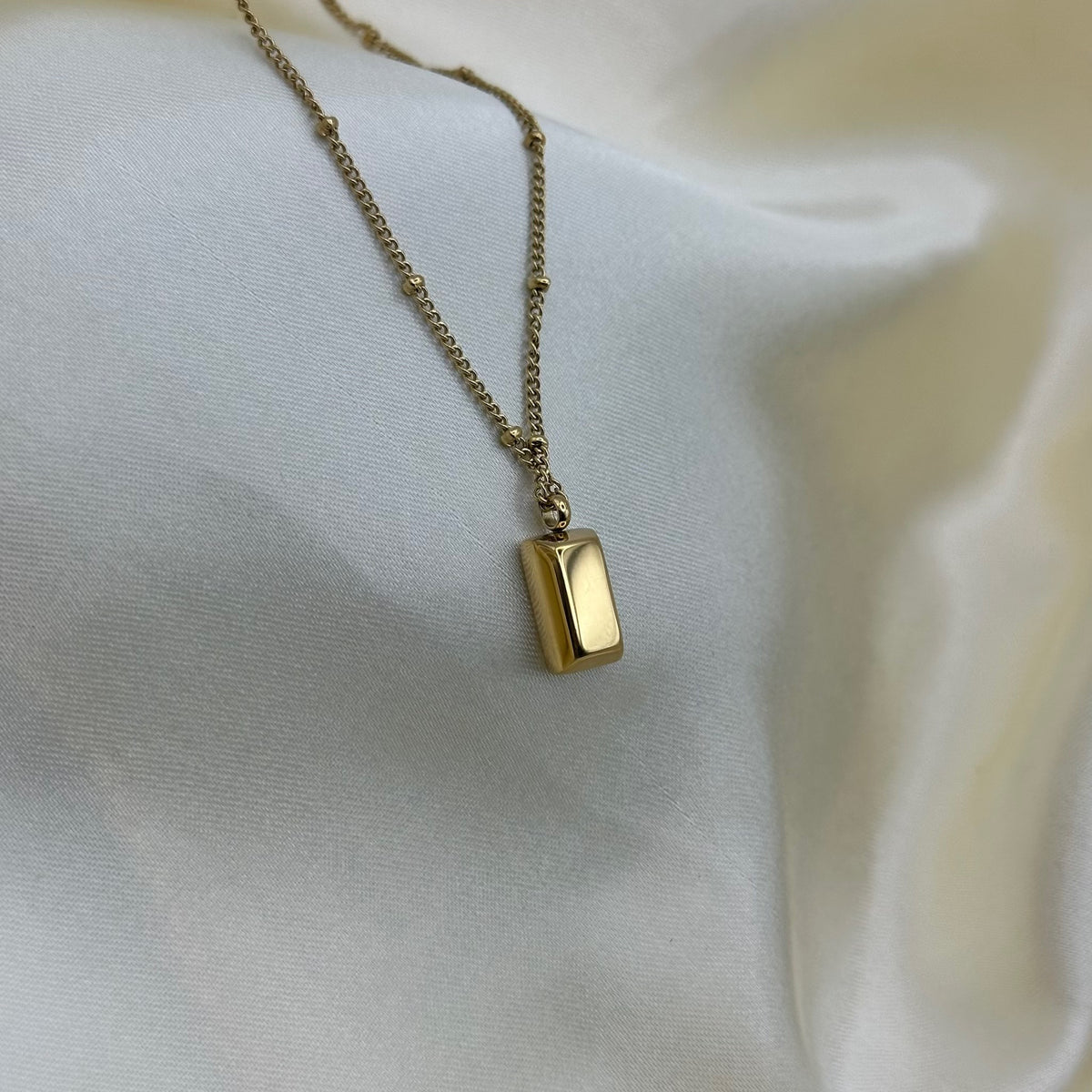 A minimalist gold ingot pendant necklace resting on soft cream satin fabric.