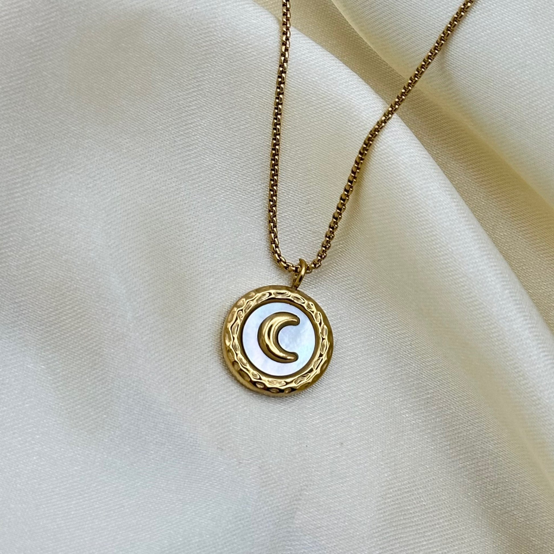 A gold crescent moon pendant set on a white enamel disc, hanging from a fine box chain on cream satin fabric.