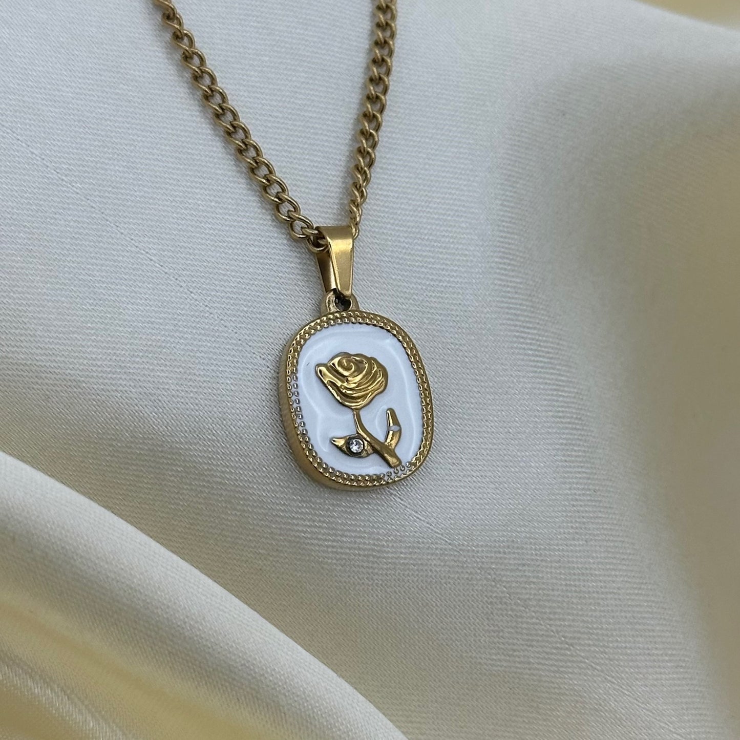 Close-up of a white enamel pendant with an embossed gold rose and small crystal, on a gold curb chain against cream fabric.