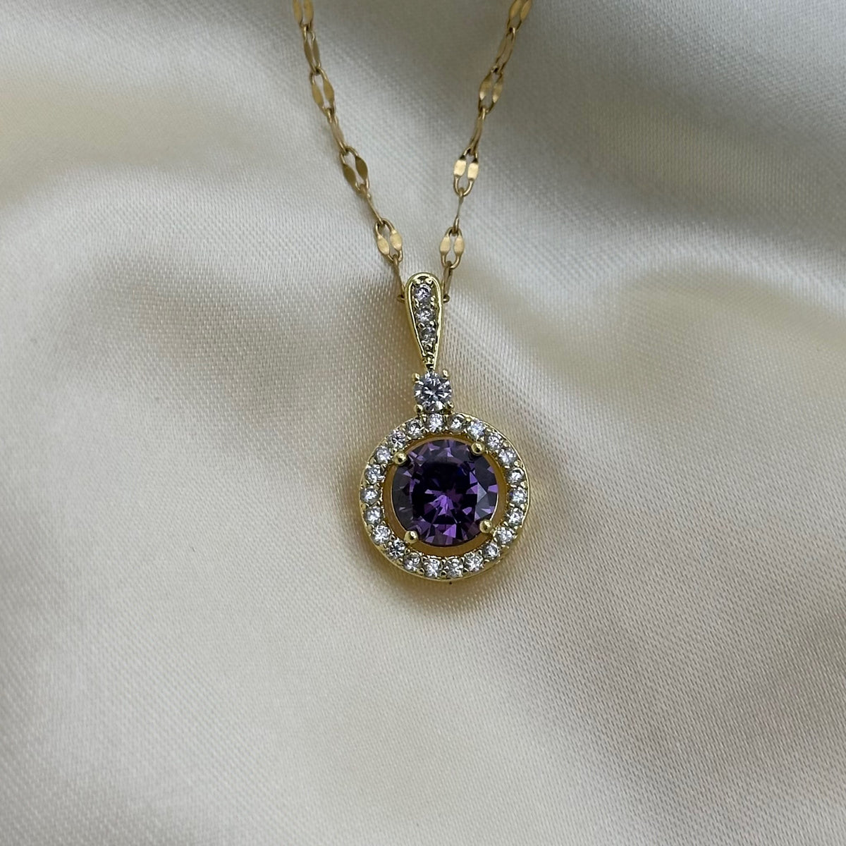 A gold pendant with a round purple crystal framed by a halo of clear stones, displayed on draped cream satin fabric.