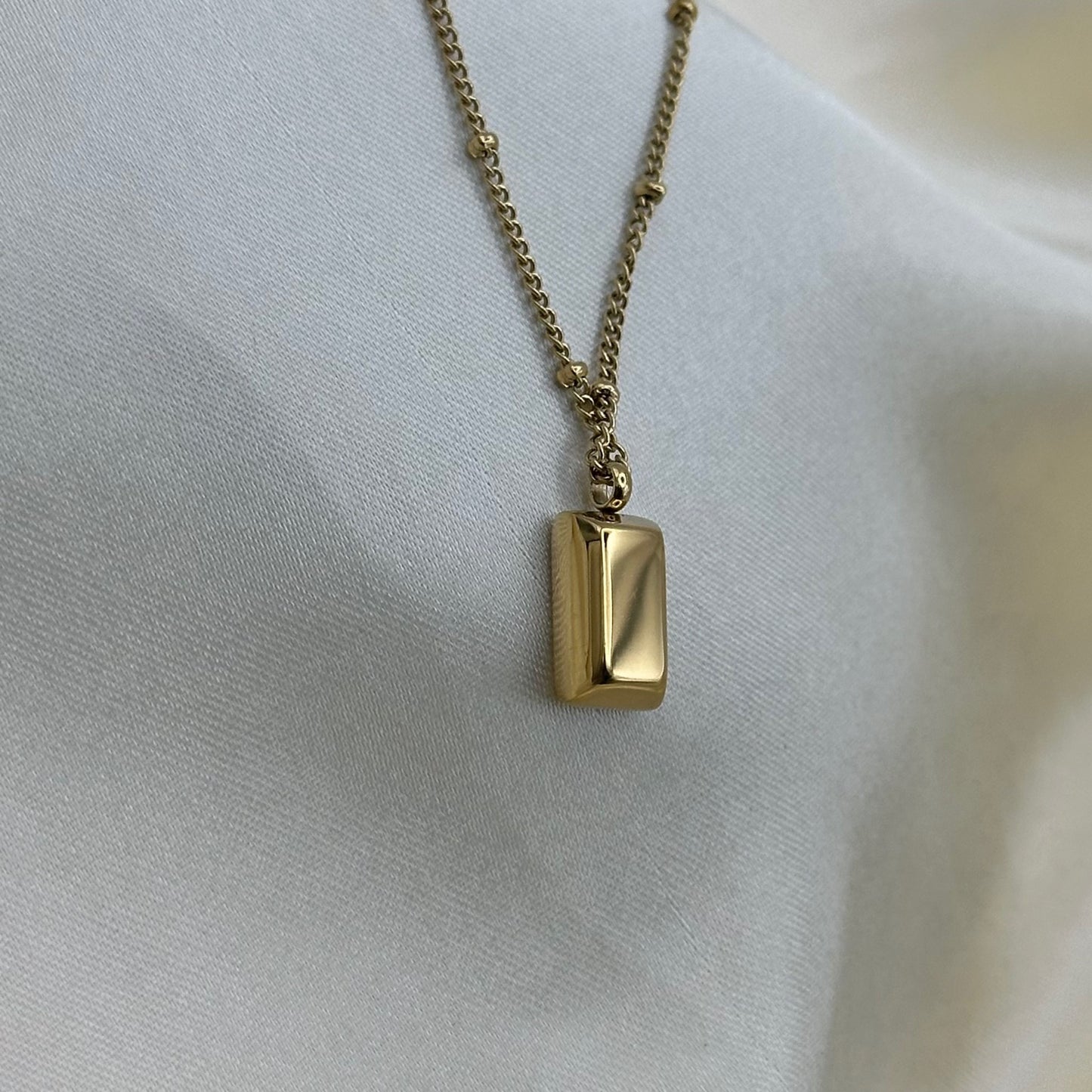 Close-up of a polished gold bar pendant on a fine beaded gold chain against cream satin.