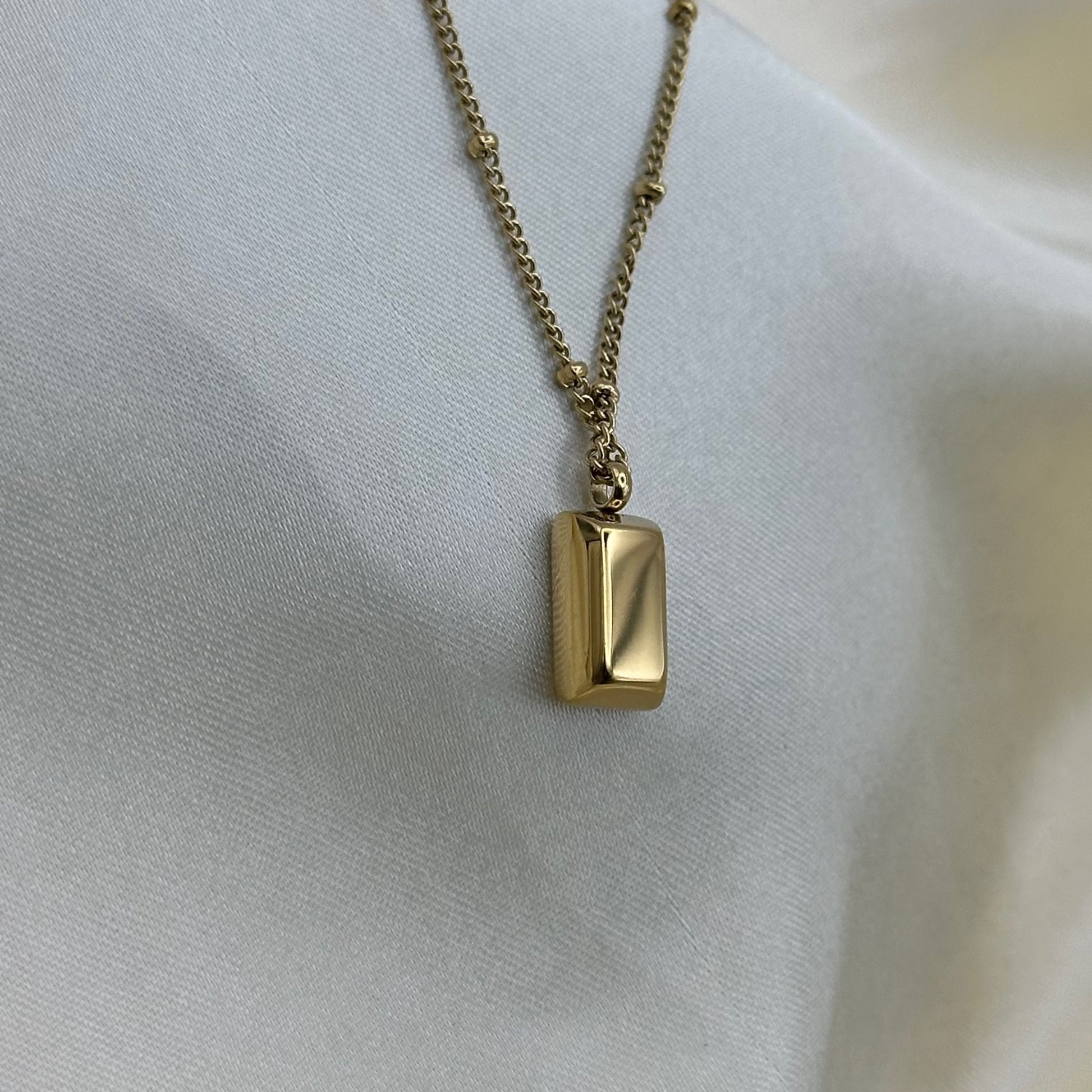 Close-up of a polished gold bar pendant on a fine beaded gold chain against cream satin.