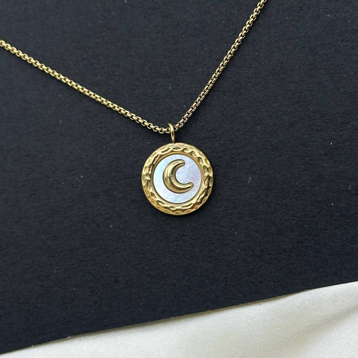 A gold circular pendant with a white mother-of-pearl face and a raised gold crescent moon, displayed on draped cream satin.