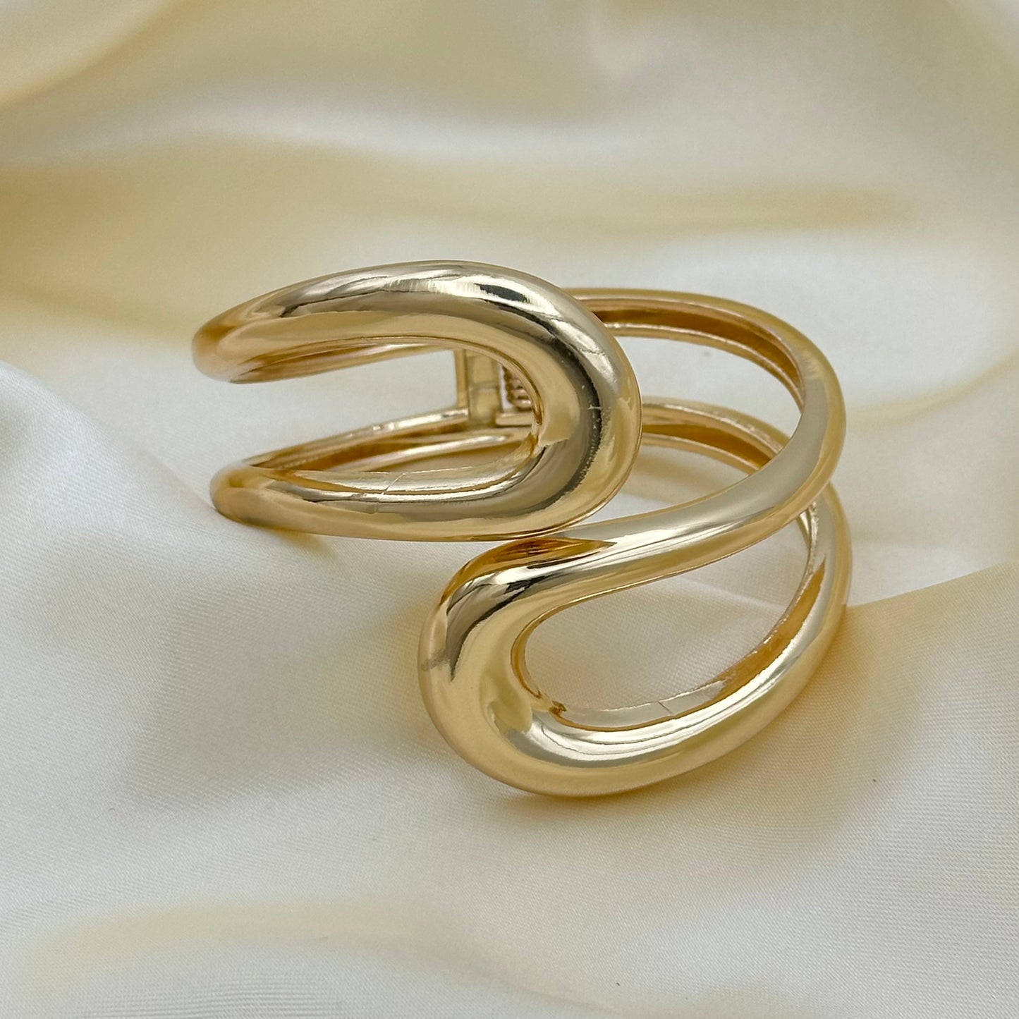 A polished gold-tone metal cuff bracelet with a twisted, open wrap design, displayed on a draped cream satin fabric background.