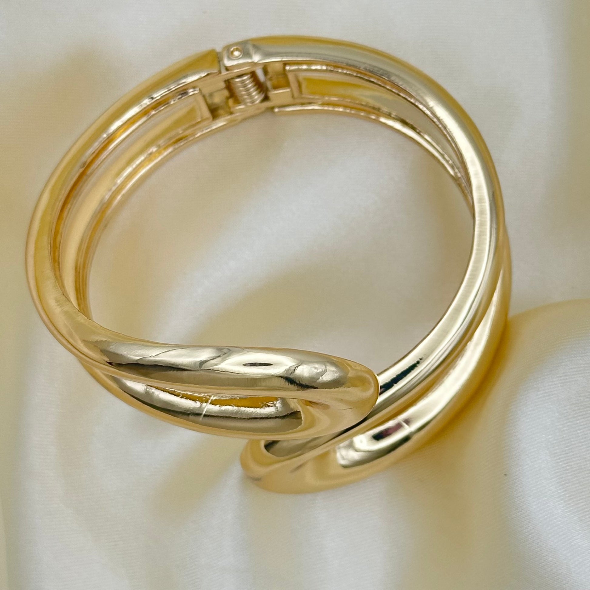 A polished gold-toned metal bangle bracelet with a twisted knot design, resting on a soft white satin fabric background.