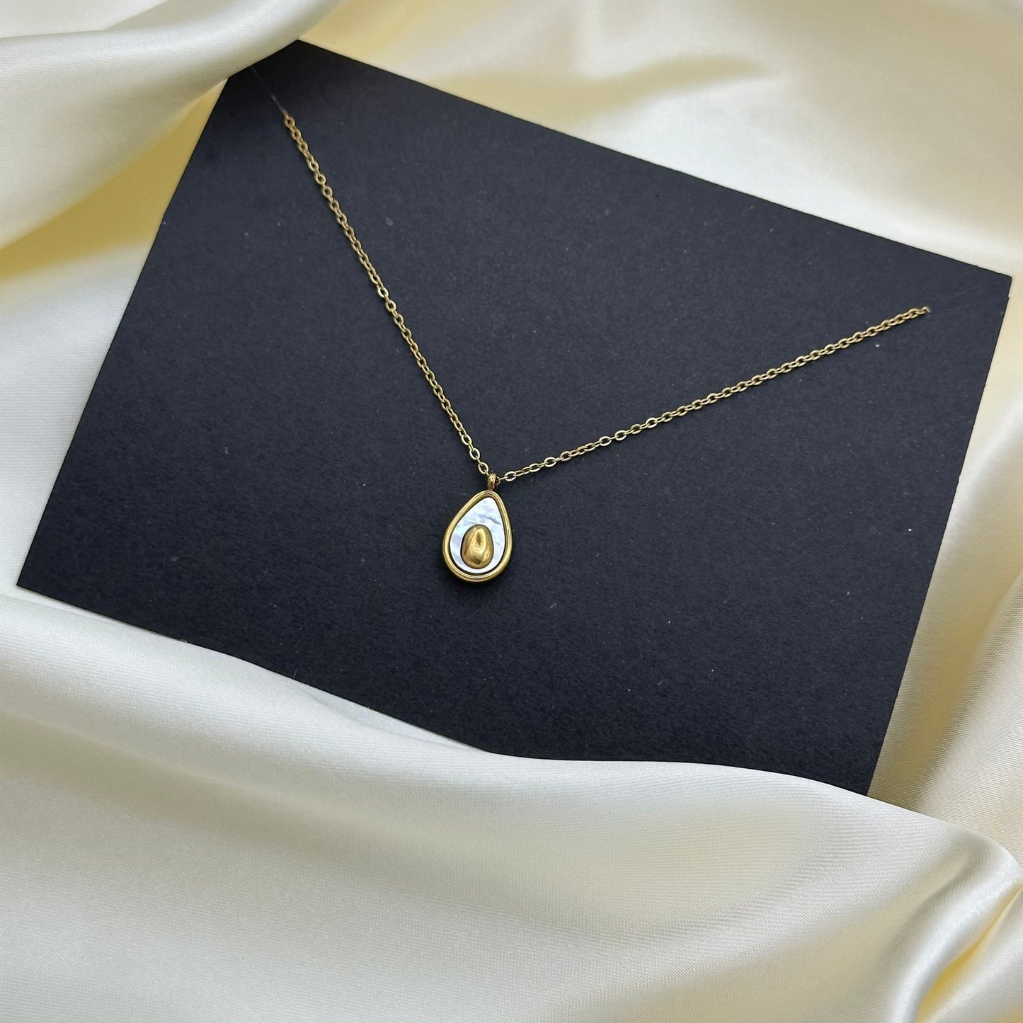A gold teardrop pendant necklace with a white inlay, displayed on a black card over draped cream satin fabric.