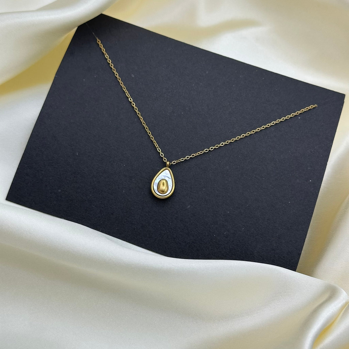 A gold teardrop pendant necklace with a white inlay, displayed on a black card over draped cream satin fabric.