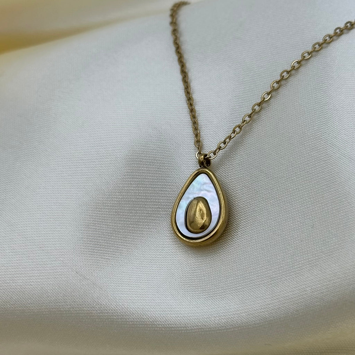 A gold chain necklace with a teardrop-shaped pendant featuring a gold center and a white iridescent shell-like outer layer, displayed on draped cream satin fabric.