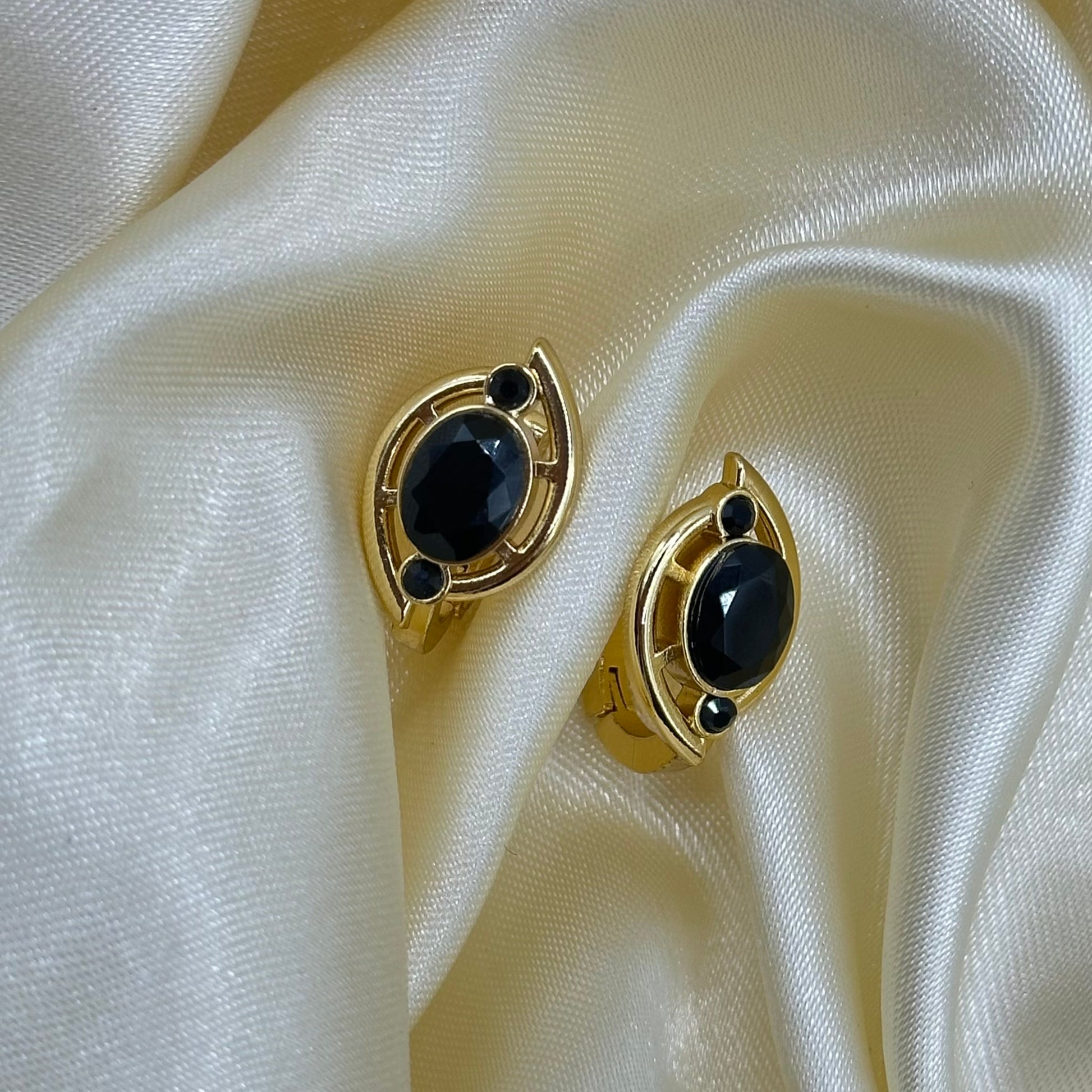 A pair of gold-toned hoop earrings with oval black faceted gemstones and small round black accent stones, displayed on draped cream satin fabric.