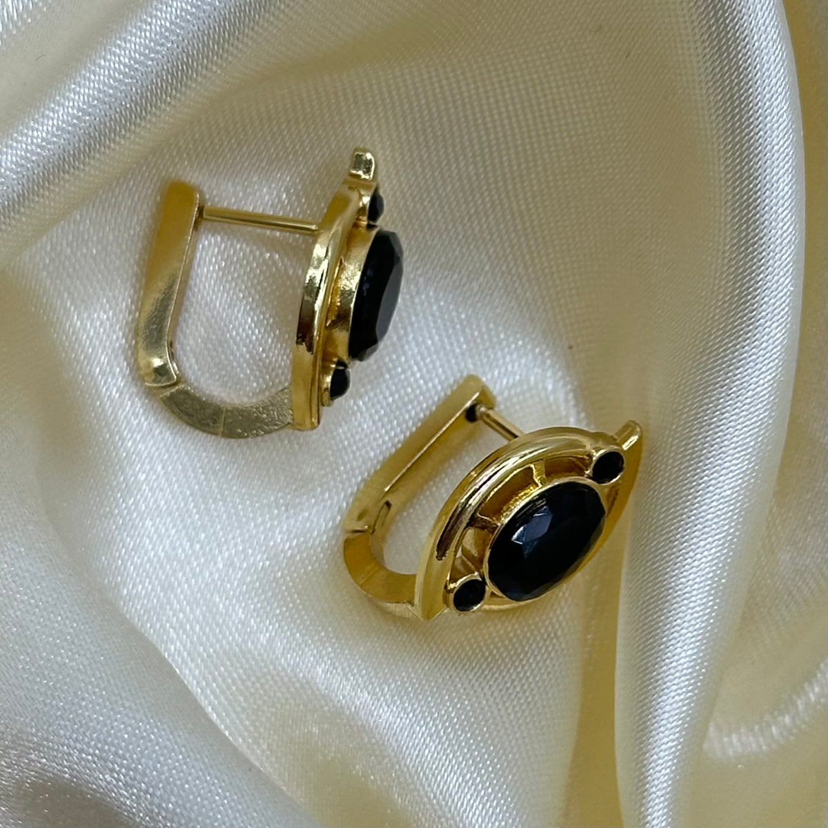 A pair of gold-toned clip-on earrings with oval black onyx stones, displayed on a draped cream satin fabric.