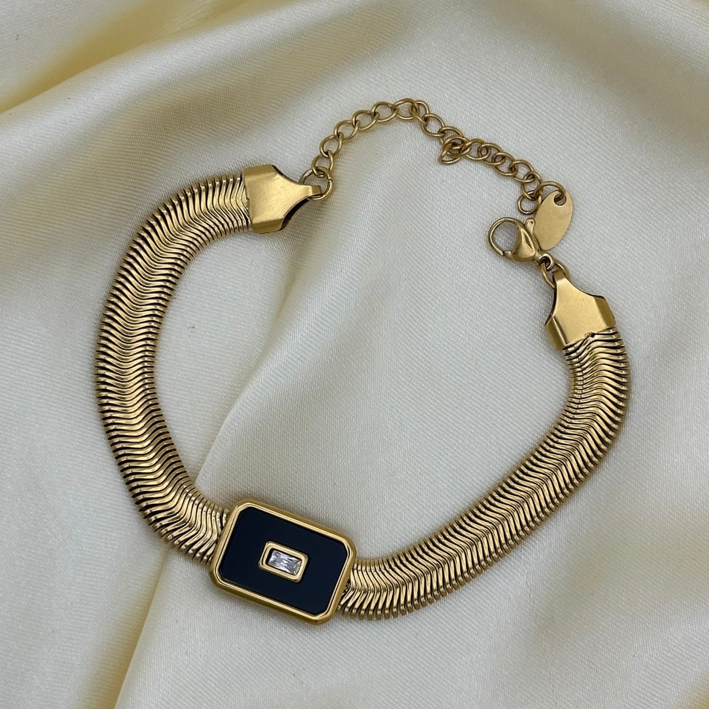 A gold-toned snake-style bracelet with a rectangular black enamel center and a small clear stone, displayed on a cream satin fabric background.