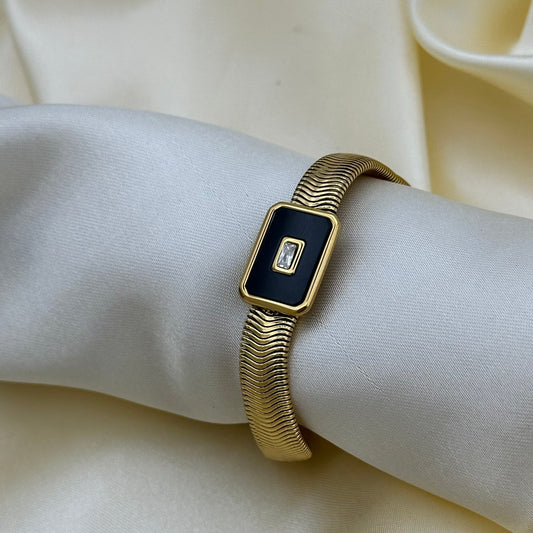 A gold-tone snake style bracelet with a rectangular black inlay and a small central crystal accent, shown worn on a cream-colored fabric wristband.