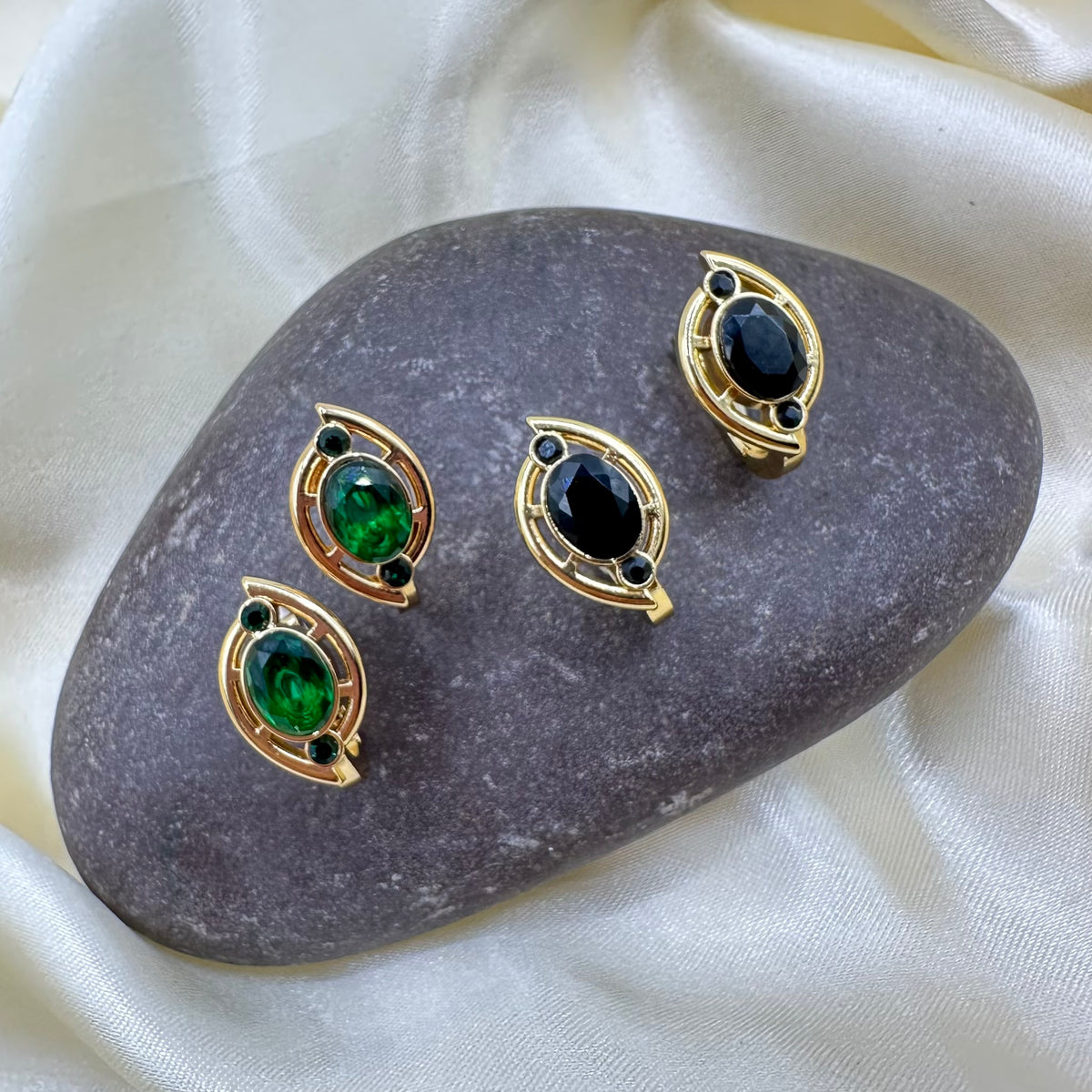 Two pairs of gold-toned earrings with oval gemstones are arranged on a smooth dark stone over cream satin fabric. Two pairs feature emerald green stones, and one pair features a black stone.