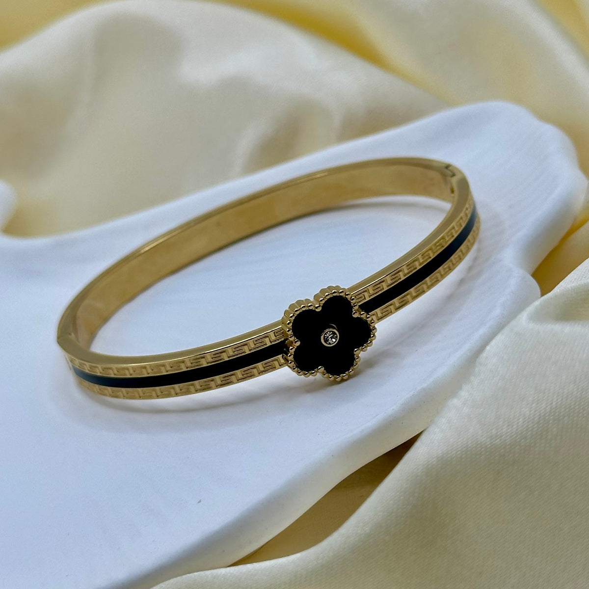 A gold-plated bangle bracelet with a black enamel flower accent and a small clear stone, displayed on a white ceramic dish over a soft cream satin fabric background.