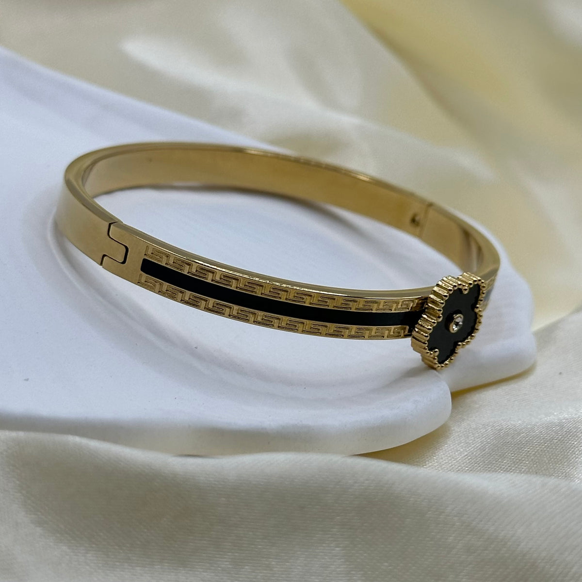 A gold-plated bangle bracelet with a black enamel center and a small clear stone, displayed on a white satin surface.