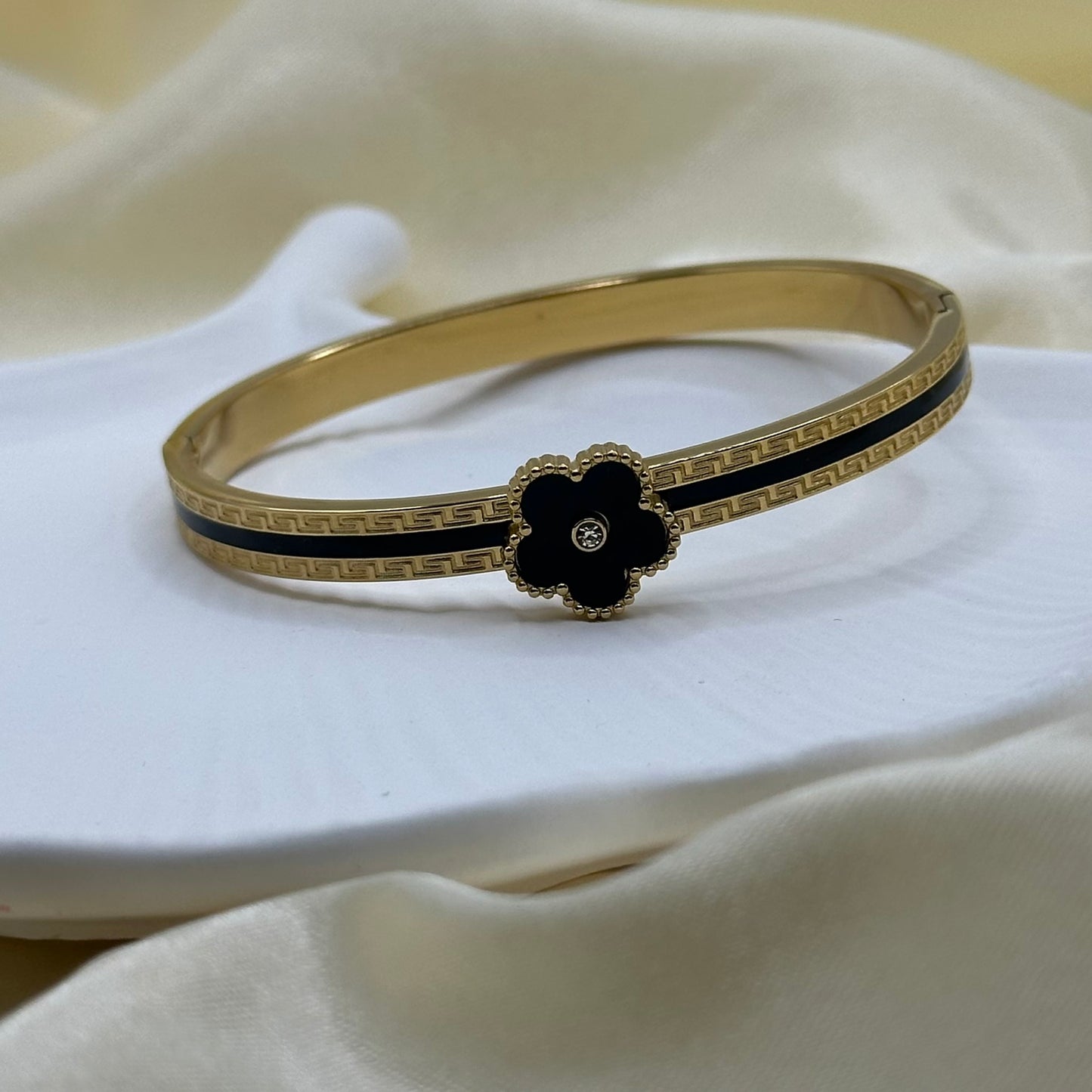 A gold-plated hinged bangle bracelet with a black enamel flower motif and Greek key detailing, resting on a white ceramic dish with draped cream-colored fabric in the background.
