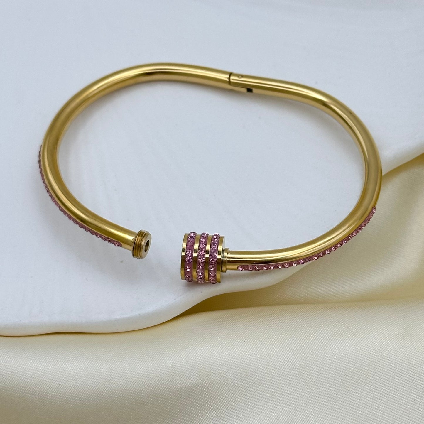 A gold-colored metal cuff bracelet with a central cluster of pink gemstones, displayed on a cream satin fabric background.
