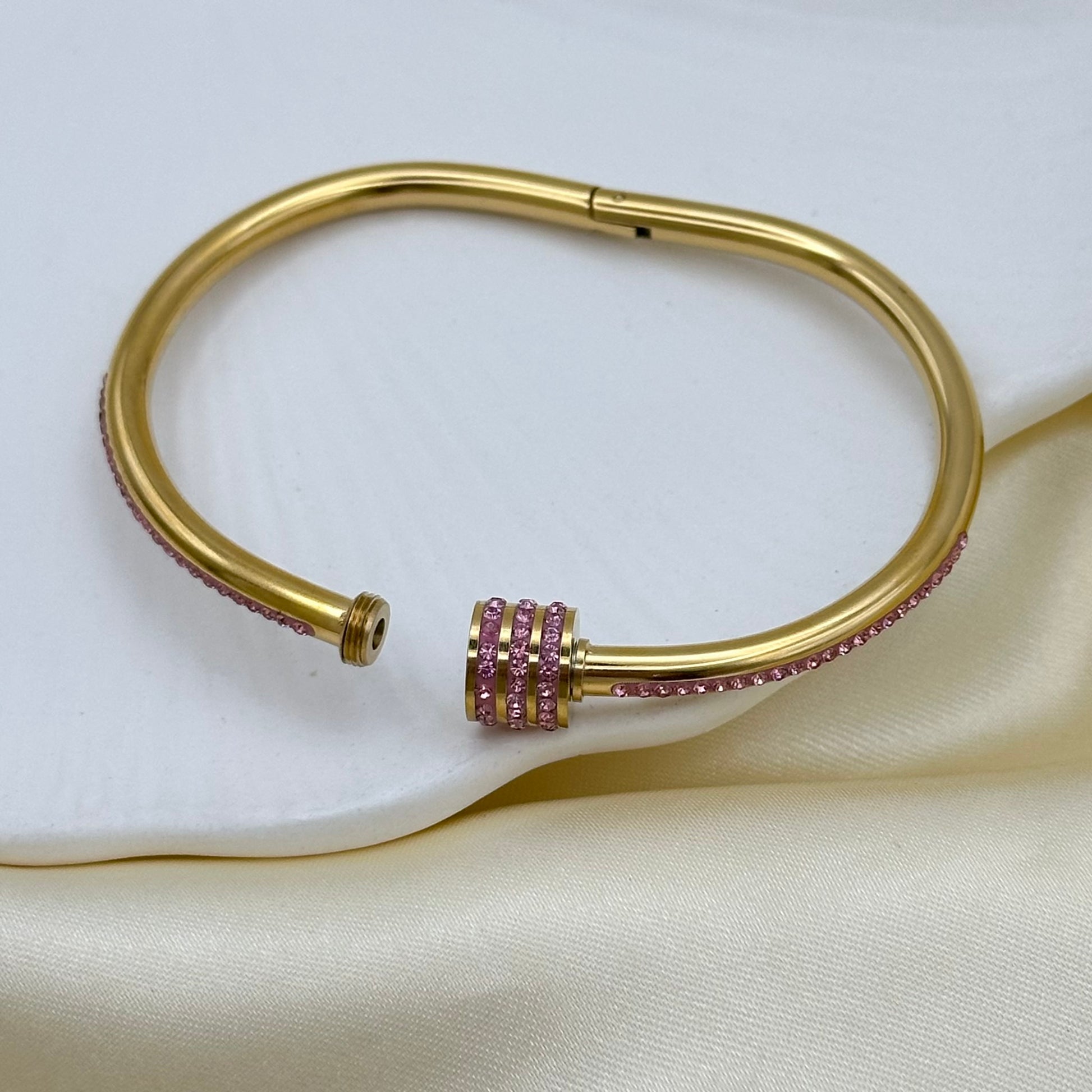 A gold-colored metal cuff bracelet with a central cluster of pink gemstones, displayed on a cream satin fabric background.