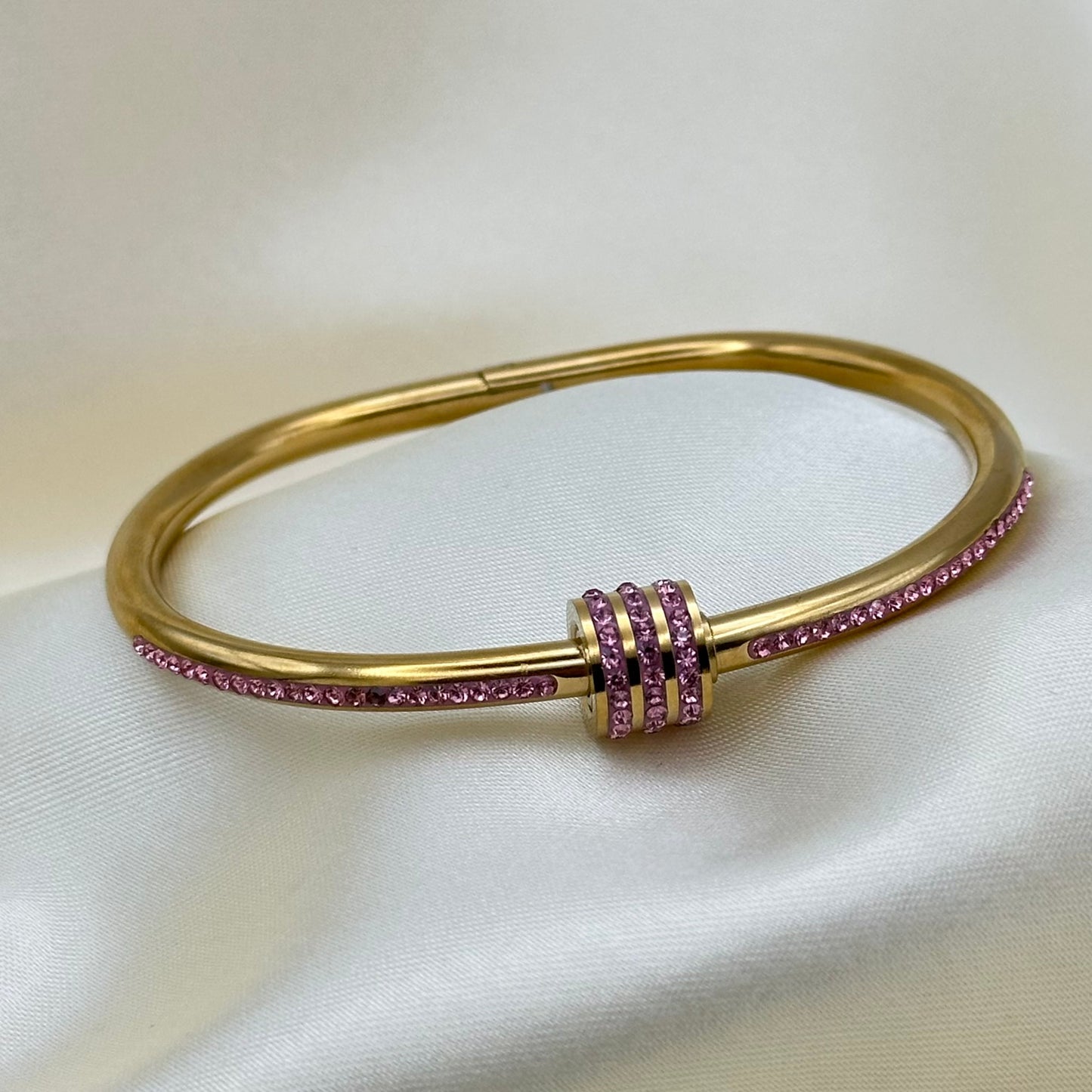 A gold-plated hinged bangle bracelet featuring rows of small pink gemstones along the outer band and a matching pink gemstone accent at the center clasp, displayed on a white satin fabric background.