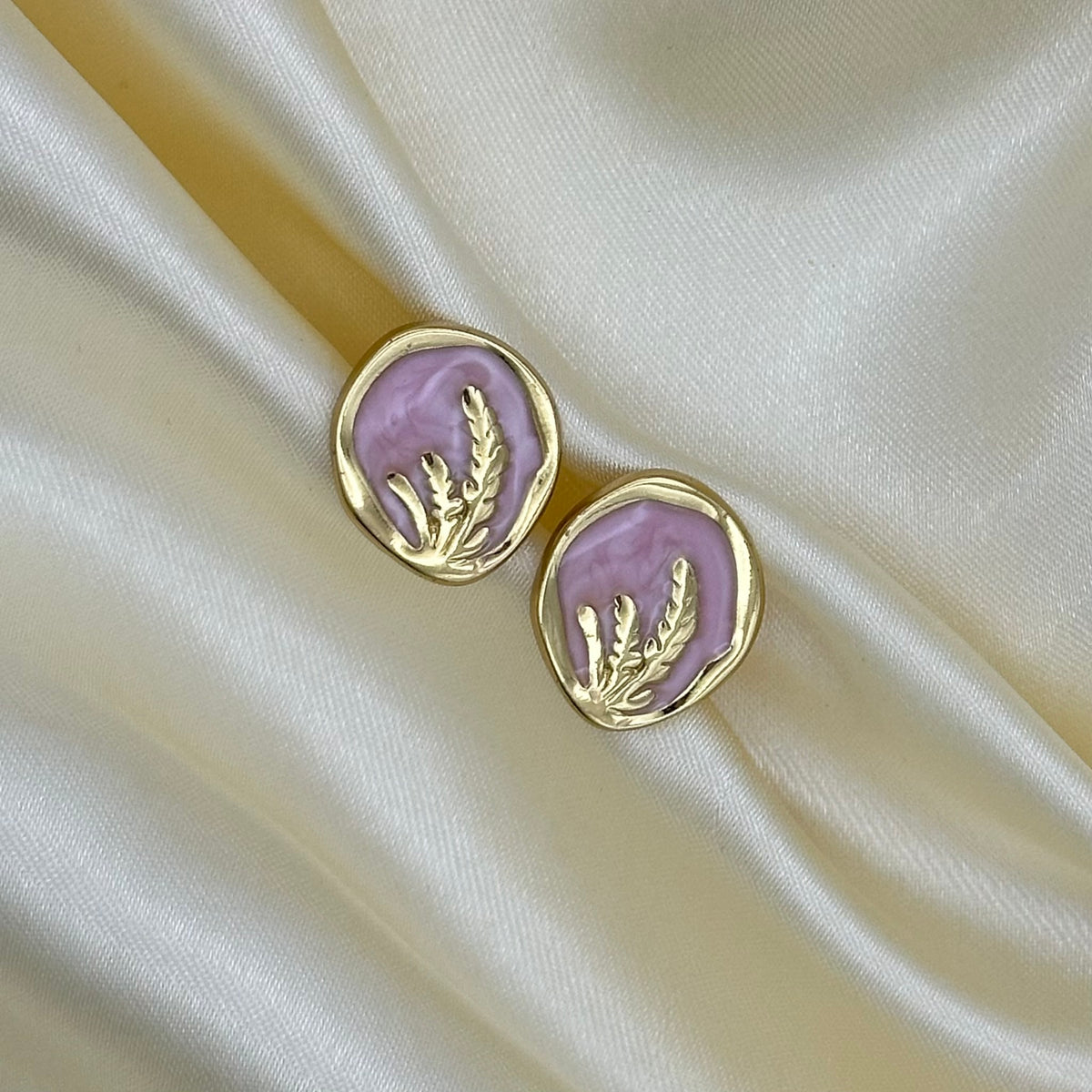 A pair of gold oval stud earrings with a light pink enamel background and a gold leaf design, displayed on draped cream satin fabric.