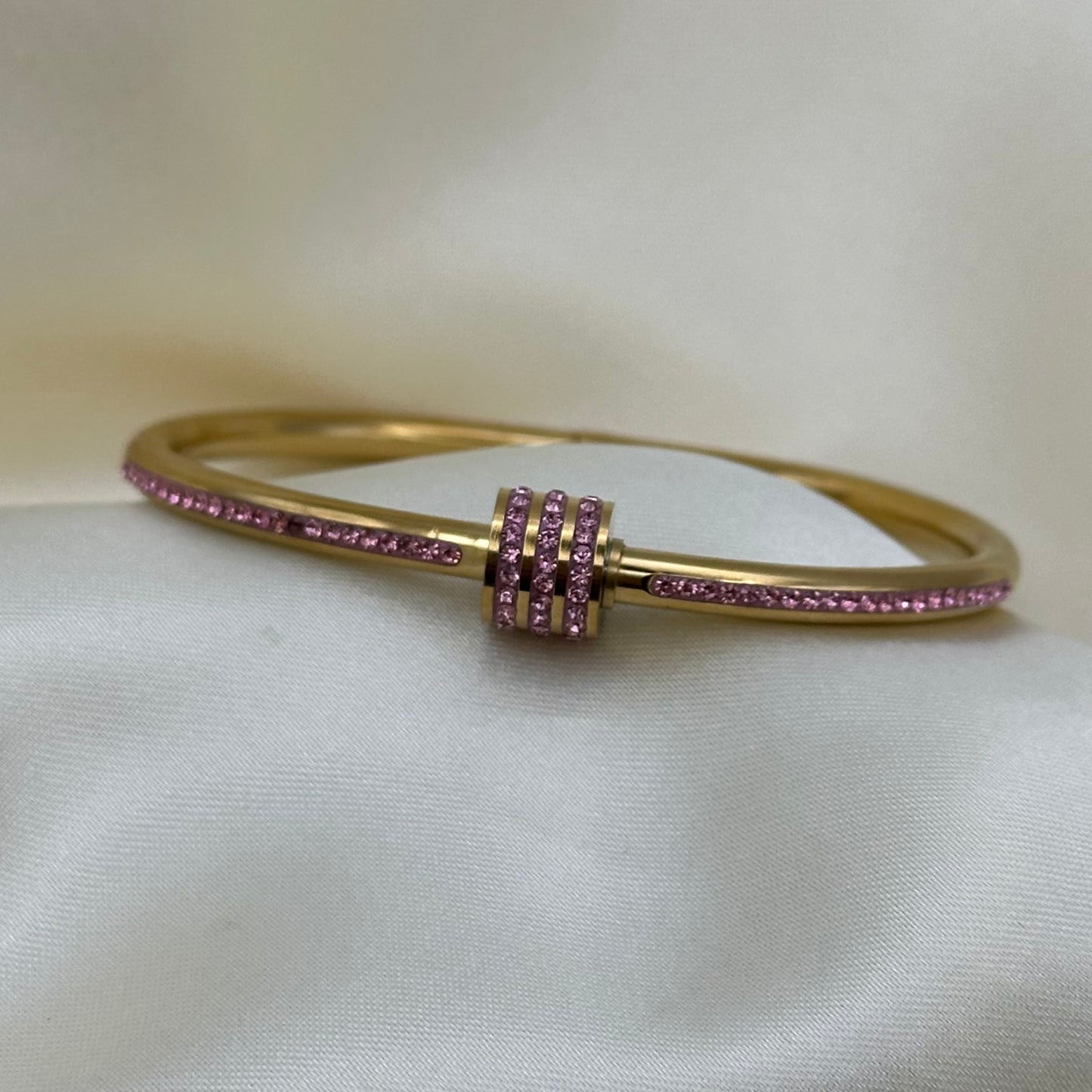 A gold bangle bracelet with rows of small pink gemstones, displayed on a soft white satin fabric background.