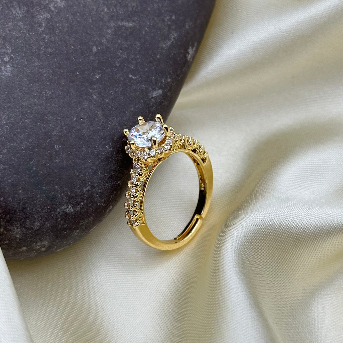 A gold ring with a large clear round-cut center stone and small clear stones along the band, styled on cream satin fabric next to a dark gray stone.