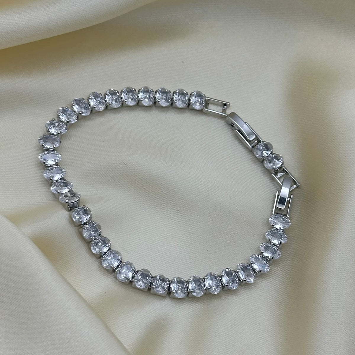 A silver-tone tennis bracelet adorned with clear round-cut crystals, displayed on a draped cream satin fabric background.
