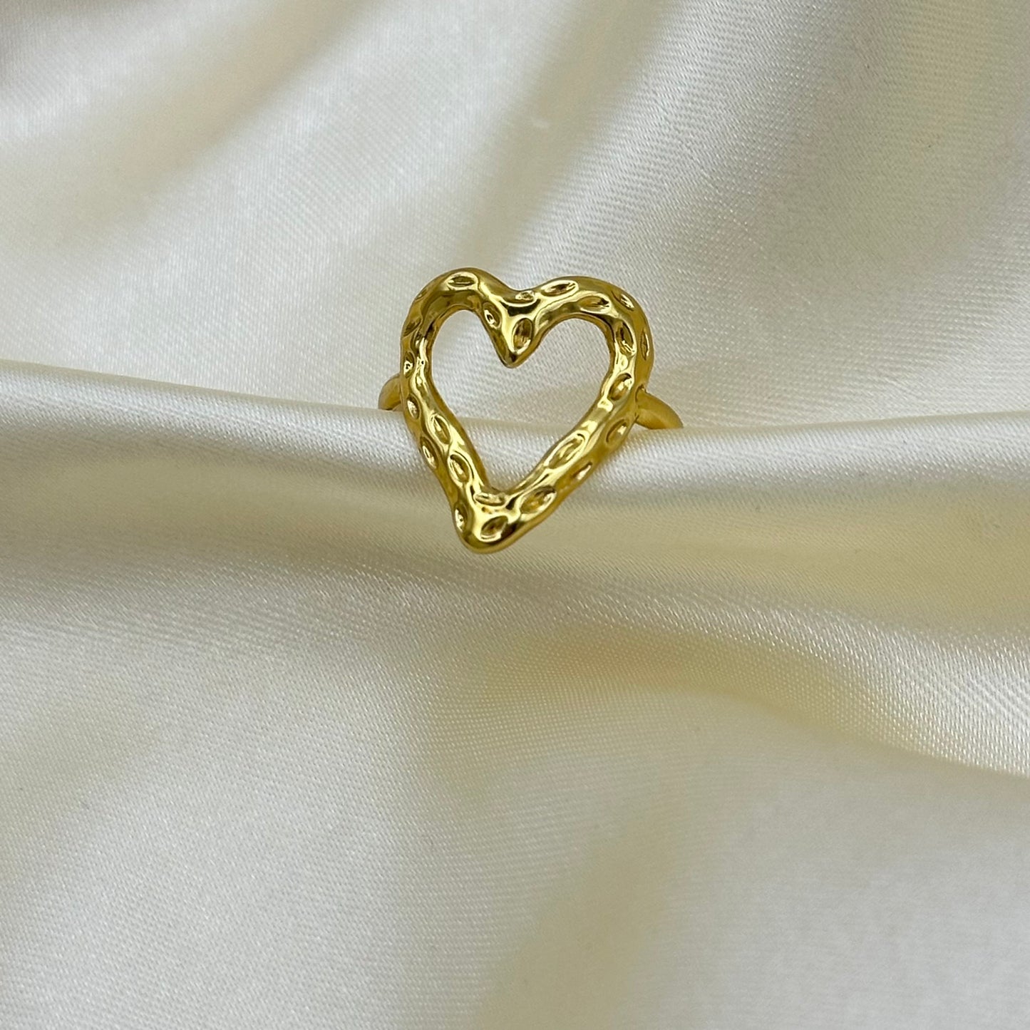 A textured gold heart-shaped ring displayed on draped cream-colored satin fabric.