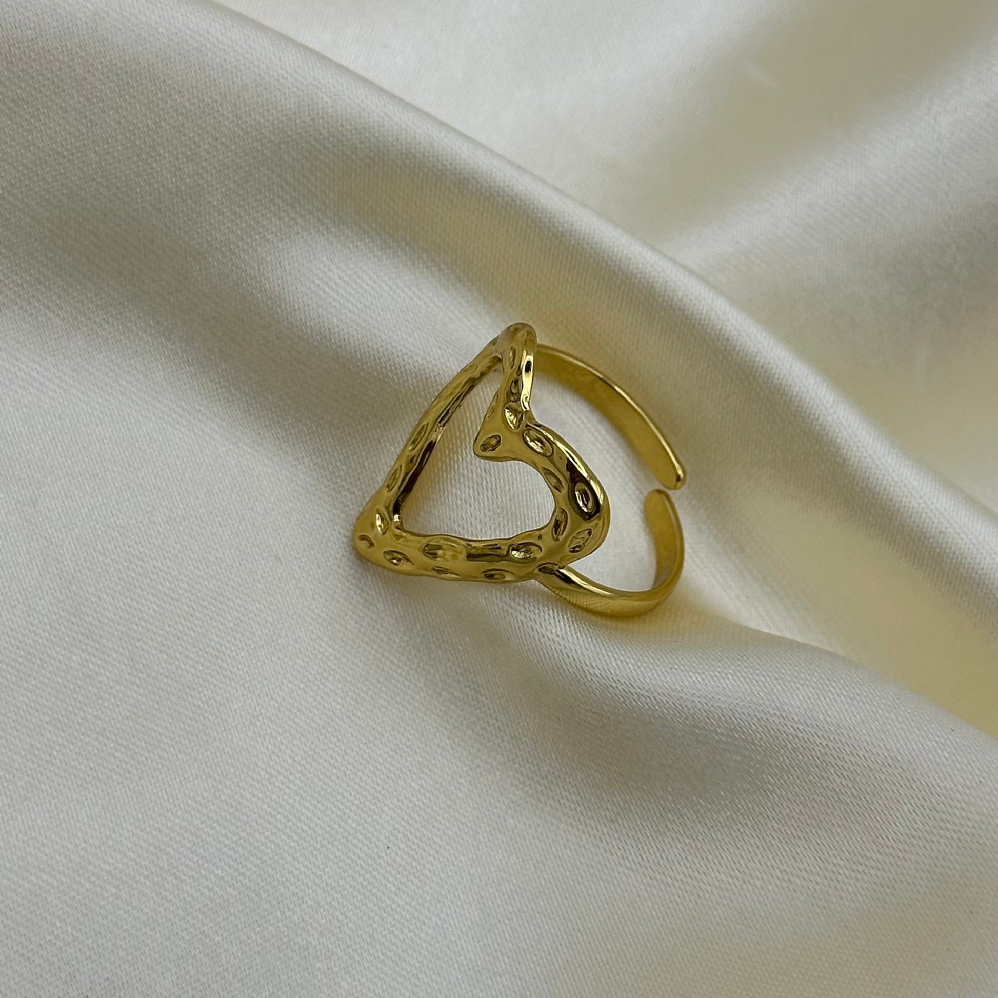A gold-toned open triangular ring with a hammered texture, displayed on draped cream-colored satin fabric.